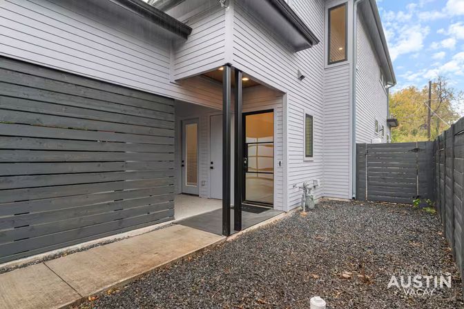 Contemporary two-story townhouse courtyard with light gray horizontal siding, covered entry supported by black metal posts, black-framed glass door, concrete walkway and gravel yard enclosed by dark horizontal slat privacy fence.