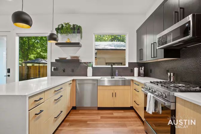 Bright modern kitchen with light wood lower cabinets and matte black uppers, white quartz countertops, stainless farmhouse sink, dishwasher and gas range, black hex-tile backsplash, pendant lights and window overlooking a leafy backyard