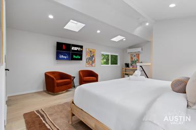 Bright modern Austin vacation rental bedroom with skylights, white king bed, two rust‑orange accent chairs, wall‑mounted TV showing streaming apps, hardwood floors and a small work nook.