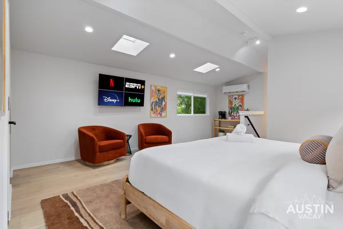 Bright modern Austin vacation rental bedroom with skylights, white king bed, two rust‑orange accent chairs, wall‑mounted TV showing streaming apps, hardwood floors and a small work nook.