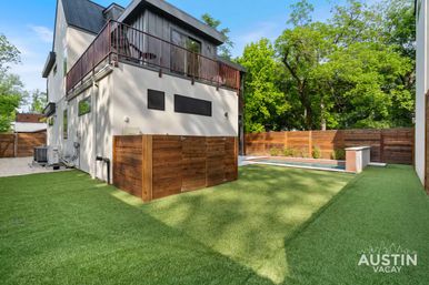 Contemporary two-story home with upper balcony overlooking a sunlit, fenced urban backyard featuring artificial turf, a narrow rectangular plunge pool, wooden privacy fencing and mature trees.