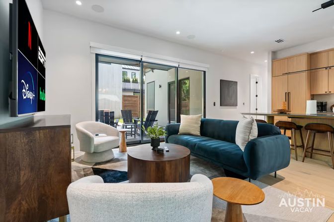 Bright modern living room with a teal velvet sofa, curved white armchairs, round wooden coffee table, wall-mounted TV showing streaming apps, sliding glass doors to a patio and an open-plan kitchen with bar stools.