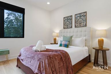 Bright modern Austin vacation rental bedroom with tufted white headboard, wooden nightstands and lamps, patterned pillows, plush mauve throw and folded towels.
