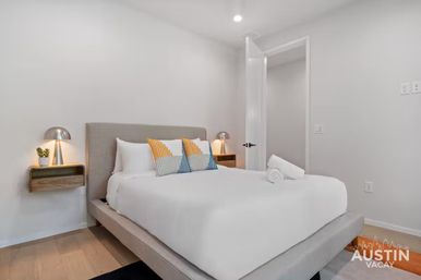 Bright modern Austin vacation rental bedroom with gray upholstered platform bed, crisp white linens, yellow-and-blue striped accent pillows, wall-mounted wooden nightstands and sleek metal lamps