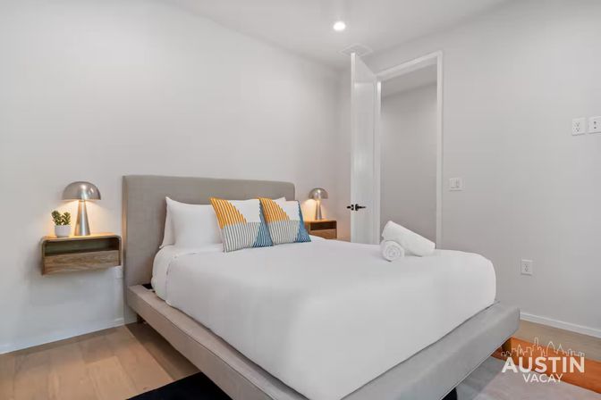 Bright modern Austin vacation rental bedroom with gray upholstered platform bed, crisp white linens, yellow-and-blue striped accent pillows, wall-mounted wooden nightstands and sleek metal lamps