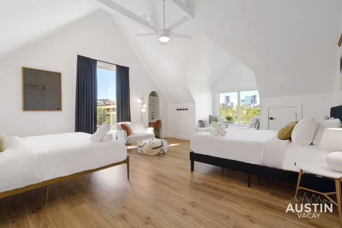 Sunlit attic-style Austin vacation rental bedroom with vaulted white ceilings, two beds in crisp white linens, hardwood floors, cozy seating nook and large windows framing the downtown skyline.