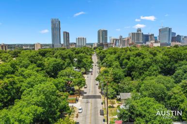 Aerial view of a tree-lined residential street leading toward the downtown Austin skyline with high-rise towers under a bright blue sky.