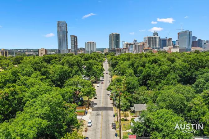 Aerial view of a tree-lined residential street leading toward the downtown Austin skyline with high-rise towers under a bright blue sky.