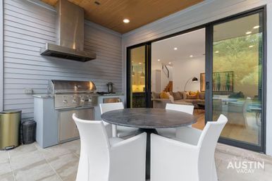 Modern Austin covered patio with stainless-steel built-in grill and hood, round black dining table with four white chairs, and sliding glass doors revealing a bright living room with beige sofa and yellow pillows.