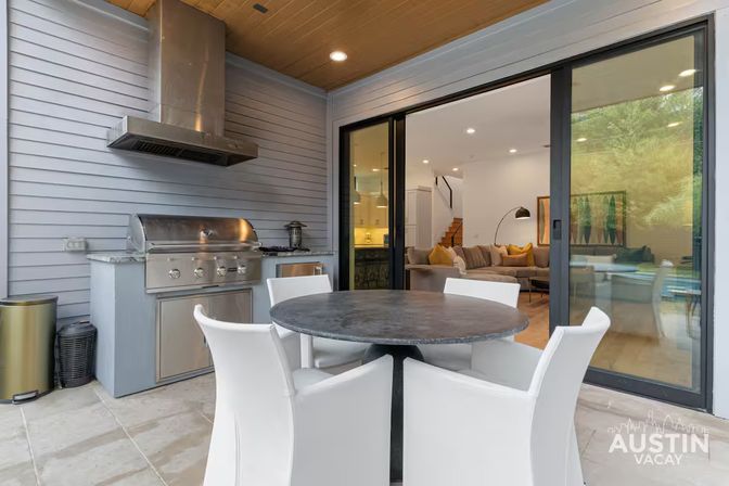 Modern Austin covered patio with stainless-steel built-in grill and hood, round black dining table with four white chairs, and sliding glass doors revealing a bright living room with beige sofa and yellow pillows.
