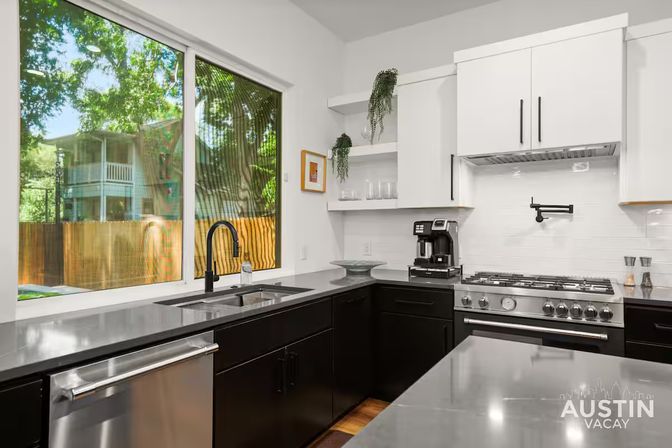 Sunny modern Austin vacation-rental kitchen with black lower cabinets, white upper cabinets, stainless appliances, sleek gray countertops, undermount sink and large window overlooking a leafy backyard.