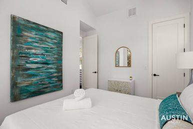 Crisp white bedroom with teal abstract wall art, rolled towel on the bed, gold arched mirror and patterned dresser — bright modern rental near Austin