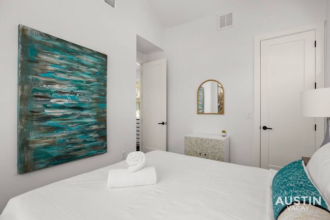 Crisp white bedroom with teal abstract wall art, rolled towel on the bed, gold arched mirror and patterned dresser — bright modern rental near Austin