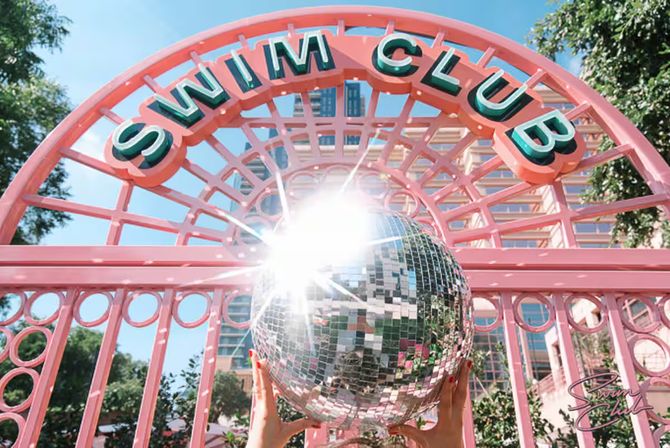 Sunlit retro pink swim club gate with teal lettering; hands lift a mirrored disco ball reflecting city buildings and sparkling light.