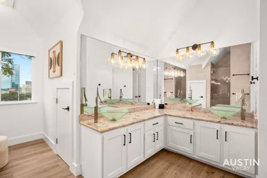Bright modern bathroom in Austin with L-shaped white vanity, marble countertop, two green glass vessel sinks, large corner mirror, exposed-bulb vanity lights, wood floors and city skyline visible through window.