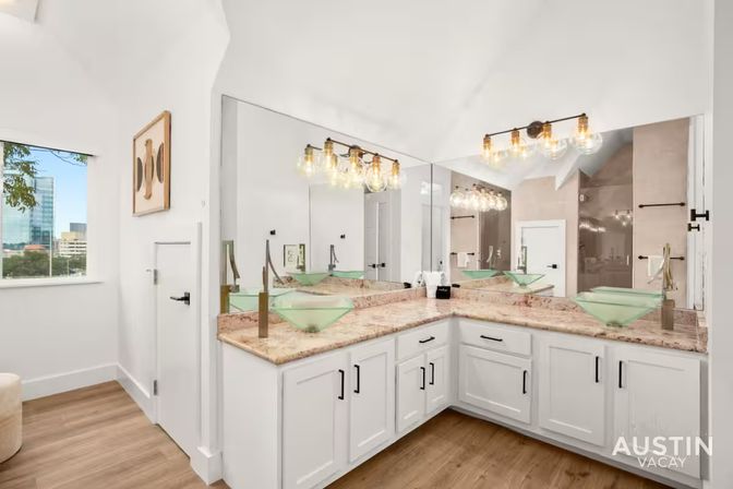 Bright modern bathroom in Austin with L-shaped white vanity, marble countertop, two green glass vessel sinks, large corner mirror, exposed-bulb vanity lights, wood floors and city skyline visible through window.