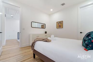 Bright modern bedroom with white walls and light hardwood floors, a neatly made bed with white linens, rolled towel and beige throw, floral accent pillow, low dresser with mirror and small plant, colorful framed art and open doorway to a hallway.