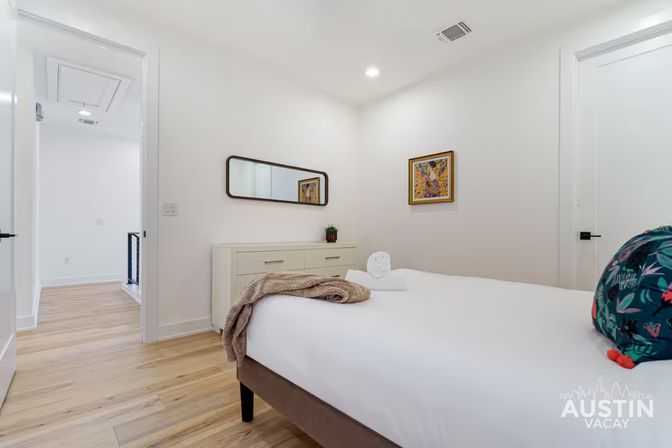 Bright modern bedroom with white walls and light hardwood floors, a neatly made bed with white linens, rolled towel and beige throw, floral accent pillow, low dresser with mirror and small plant, colorful framed art and open doorway to a hallway.