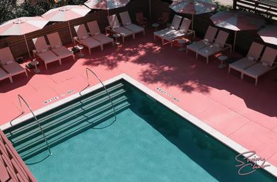 Aerial view of a turquoise outdoor pool with metal handrails and steps, set in a pink sun deck lined with lounge chairs, umbrellas and small tables.
