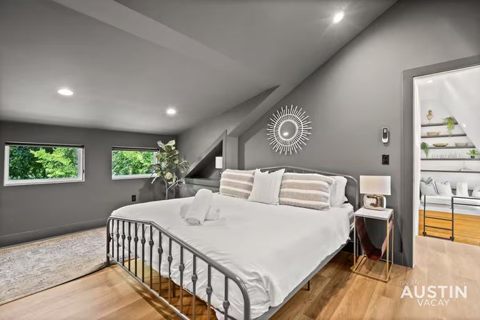 Cozy modern attic bedroom with sloped ceiling, metal-frame king bed in white linens, sunburst wall mirror, bedside lamp, hardwood floors and two small windows with leafy treetop views.