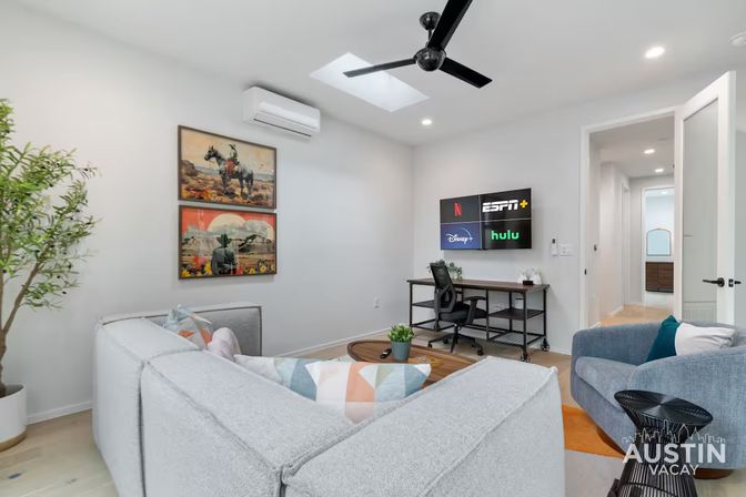 Bright modern living room with light-gray sectional and colorful pillows, blue armchair, round coffee table, wall-mounted TV showing streaming app logos, a desk with office chair, skylight and black ceiling fan, framed western art and potted plant.