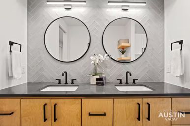 Sleek modern double-sink bathroom vanity with matte black faucets and round mirrors, gray herringbone tile backsplash, light wood cabinets, white orchid and soap dispenser — contemporary Austin rental bathroom.