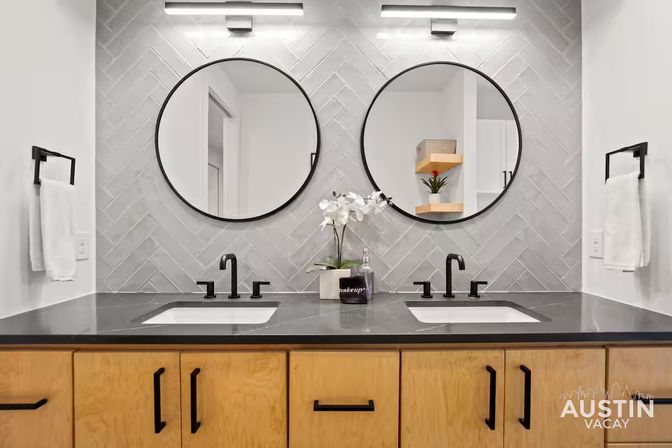 Sleek modern double-sink bathroom vanity with matte black faucets and round mirrors, gray herringbone tile backsplash, light wood cabinets, white orchid and soap dispenser — contemporary Austin rental bathroom.