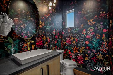 Vibrant modern powder room with dark botanical wallpaper of colorful flowers and birds, round brass mirror, rectangular white vessel sink on a dark stone countertop, wood vanity, toilet and a small window