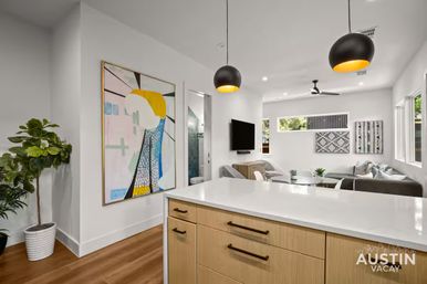 Sunlit modern Austin, TX open-plan living area with white kitchen island and wood cabinets, black-and-gold pendant lights, abstract wall art, potted fiddle-leaf fig, and cozy gray sofa seating.