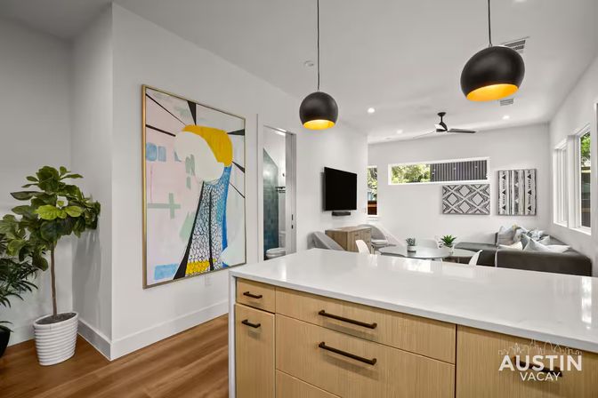 Sunlit modern Austin, TX open-plan living area with white kitchen island and wood cabinets, black-and-gold pendant lights, abstract wall art, potted fiddle-leaf fig, and cozy gray sofa seating.
