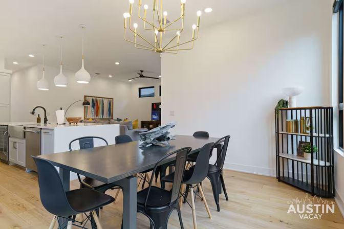 Airy Austin open-plan dining area with a sleek matte-black table and mixed metal chairs under a gold chandelier, white kitchen island with pendant lights, light wood floors, minimalist shelving and colorful wall art.