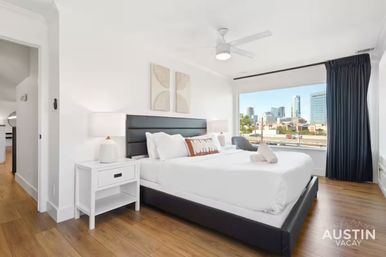 Bright, modern bedroom — king bed with black headboard, crisp white linens, bedside lamps, wood floors and large window with a downtown city skyline view.