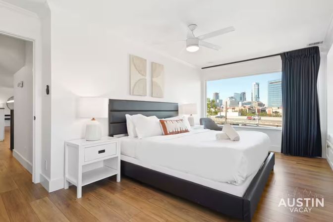 Bright, modern bedroom — king bed with black headboard, crisp white linens, bedside lamps, wood floors and large window with a downtown city skyline view.