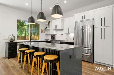 Sleek modern kitchen with a large gray island, four wooden bar stools, three black pendant lights, stainless steel refrigerator, white cabinets, hardwood floors and a window showing green trees.