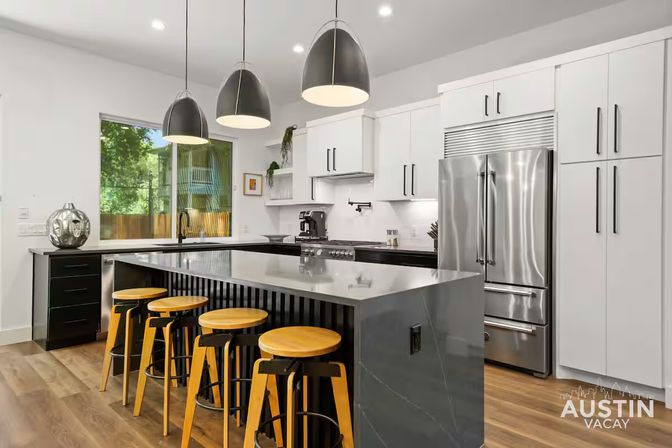 Sleek modern kitchen with a large gray island, four wooden bar stools, three black pendant lights, stainless steel refrigerator, white cabinets, hardwood floors and a window showing green trees.