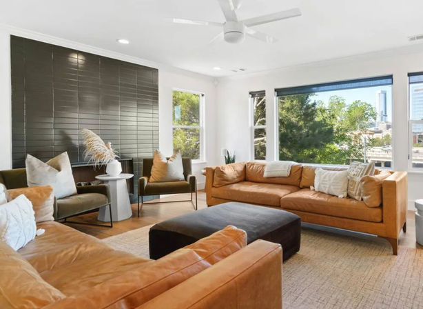 Sunlit modern living room with tan leather sofas, large dark ottoman, green accent chairs, textured rug, black tiled feature wall, ceiling fan and wide windows framing trees and an urban skyline
