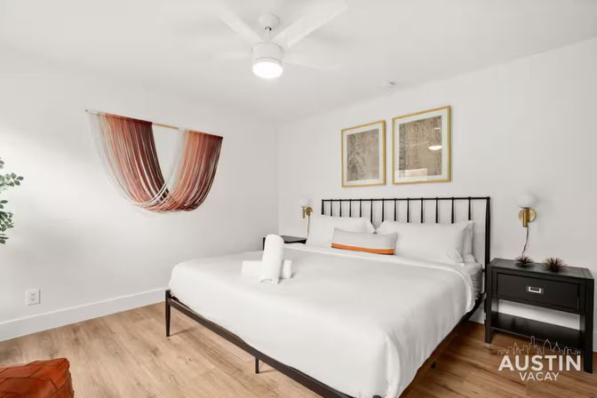 Bright modern bedroom with king bed dressed in white linens, black metal headboard, wood floors, colorful woven wall hanging, two nightstands with brass sconces and a ceiling fan — Austin vacation rental.