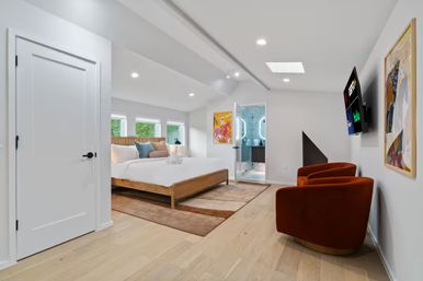 Bright modern bedroom with wooden platform bed, skylight and three windows, light hardwood floors, rust‑orange lounge chairs, wall TV and open en‑suite bathroom with blue mosaic tile