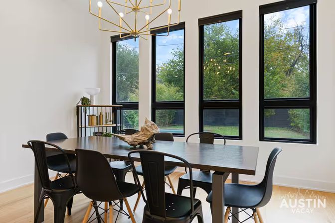Austin modern dining room with long wooden table, mixed black metal and molded chairs, geometric gold chandelier, driftwood centerpiece, and tall black‑framed windows overlooking a green backyard.