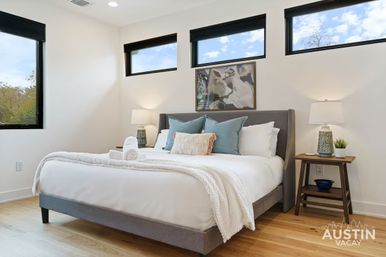 Sunny modern Austin vacation rental bedroom with a neatly made bed in white linens, blue accent pillows and rolled towels, wooden nightstands with lamps, wall art, hardwood floors, and narrow high windows.