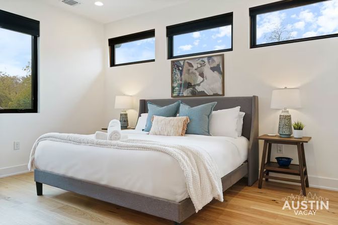 Sunny modern Austin vacation rental bedroom with a neatly made bed in white linens, blue accent pillows and rolled towels, wooden nightstands with lamps, wall art, hardwood floors, and narrow high windows.