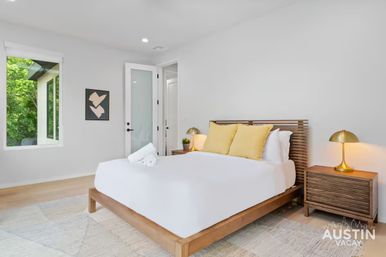 Bright, modern minimalist bedroom with wooden platform bed, crisp white linens and yellow accent pillows, matching wood nightstands with brass lamps, rolled towels and a large window framing green trees — cozy vacation rental interior.