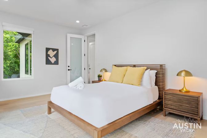Bright, modern minimalist bedroom with wooden platform bed, crisp white linens and yellow accent pillows, matching wood nightstands with brass lamps, rolled towels and a large window framing green trees — cozy vacation rental interior.