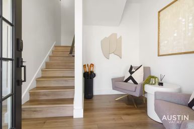 Bright modern entryway with light wood stairs, umbrella stand, gray mid-century armchairs with black-and-white pillows, round white side table, abstract wall art and warm hardwood floors.