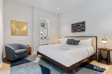 Bright modern bedroom in Austin with a low-profile wood platform bed, crisp white linens and navy accent pillows, gold bedside lamps, abstract wall art, blue geometric rug, gray armchair and a doorway to an ensuite bathroom.