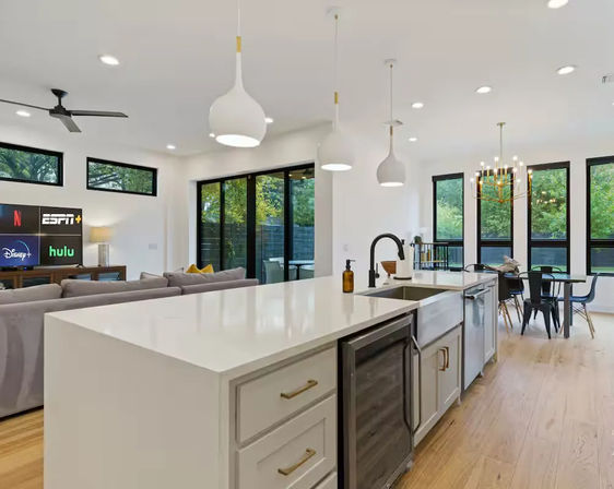 Modern open-concept kitchen and living room with large white island and quartz countertop, matte black faucet, pendant lights, hardwood floors, black-framed windows and sliding glass doors overlooking a green yard, dining table and sofa with TV showing streaming apps.