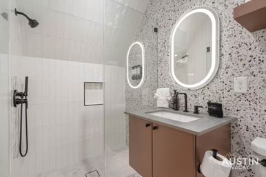 Chic modern bathroom with glass walk-in shower and matte black fixtures, terrazzo accent wall, illuminated oval vanity mirror, concrete countertop and brown cabinet.