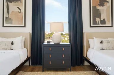 Modern twin bedroom in an Austin vacation rental — two white beds flank a navy three-drawer dresser topped with a textured lamp, framed abstract art, and floor-to-ceiling navy curtains.