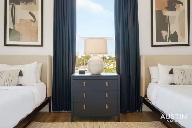 Modern twin bedroom in an Austin vacation rental — two white beds flank a navy three-drawer dresser topped with a textured lamp, framed abstract art, and floor-to-ceiling navy curtains.