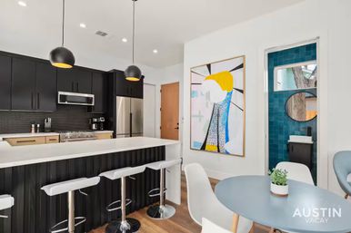 Modern Austin kitchen-dining area with black cabinets, white island, chrome bar stools, pendant lights, stainless appliances, bold abstract wall art and teal-tiled bathroom doorway.